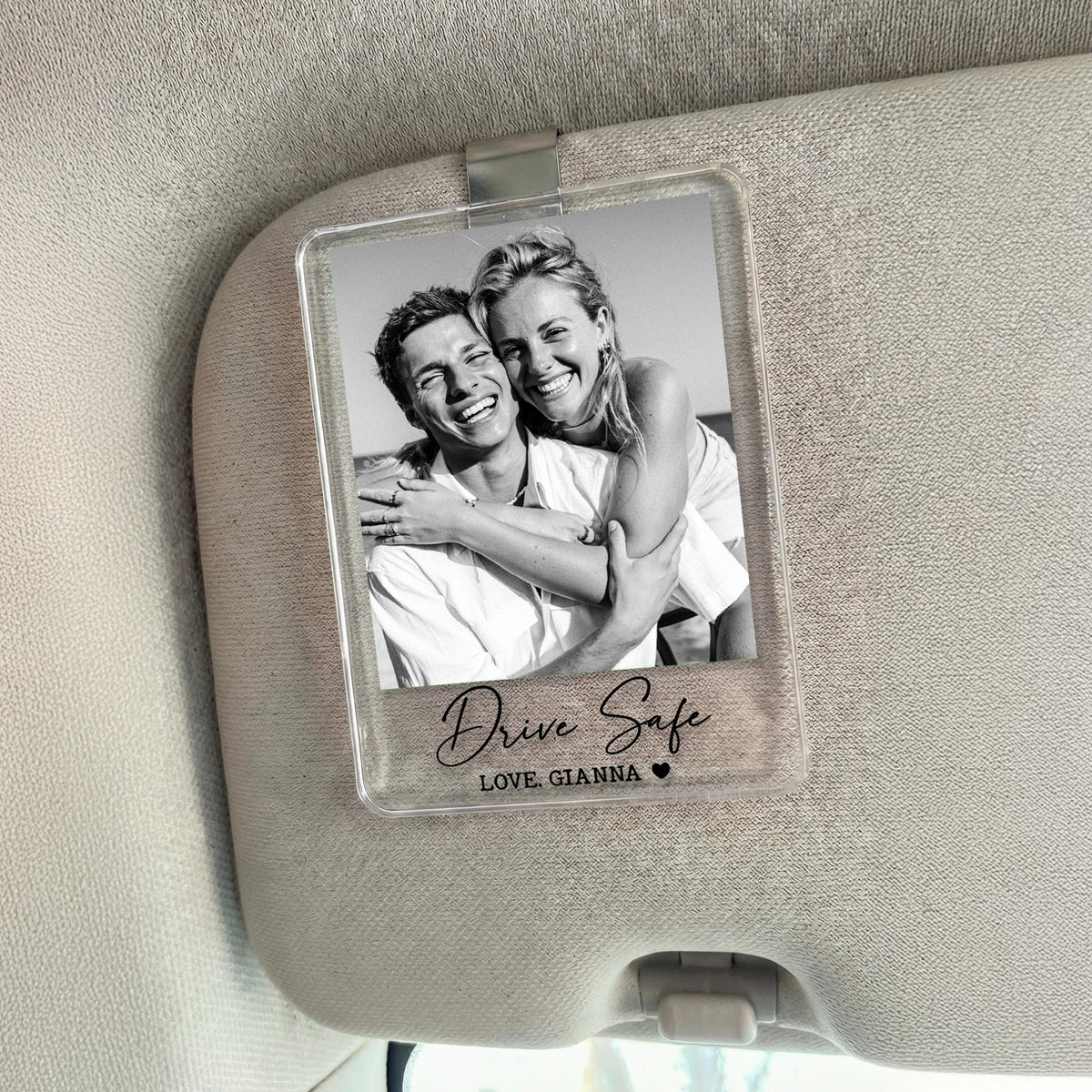 A clear acrylic car visor clip featuring a black and white photo of a smiling couple. The text "Drive Safe" and "LOVE, GIANNA" are printed on the clip, along with a small heart symbol.