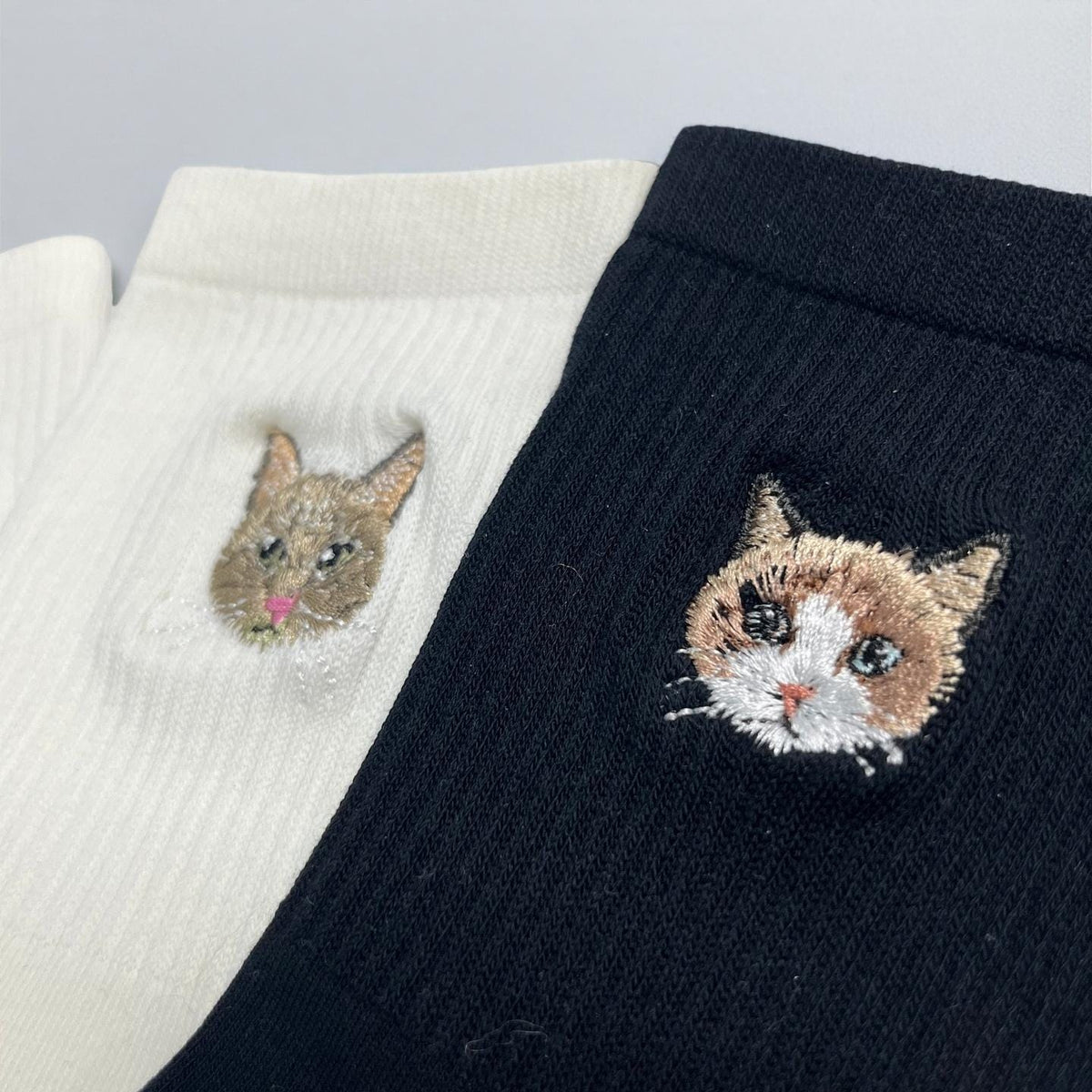 A pair of ribbed socks, one in white and one in black, each featuring a detailed embroidered cat face. The white sock displays a brown cat with a pink nose, while the black sock shows a calico cat with white, brown, and black fur.