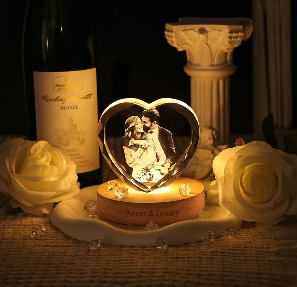 A heart-shaped glass block featuring a laser-etched photograph of a couple, illuminated by a round wooden base with the engraved text Peter & Jenny. The display is surrounded by white artificial roses and small clear crystals.