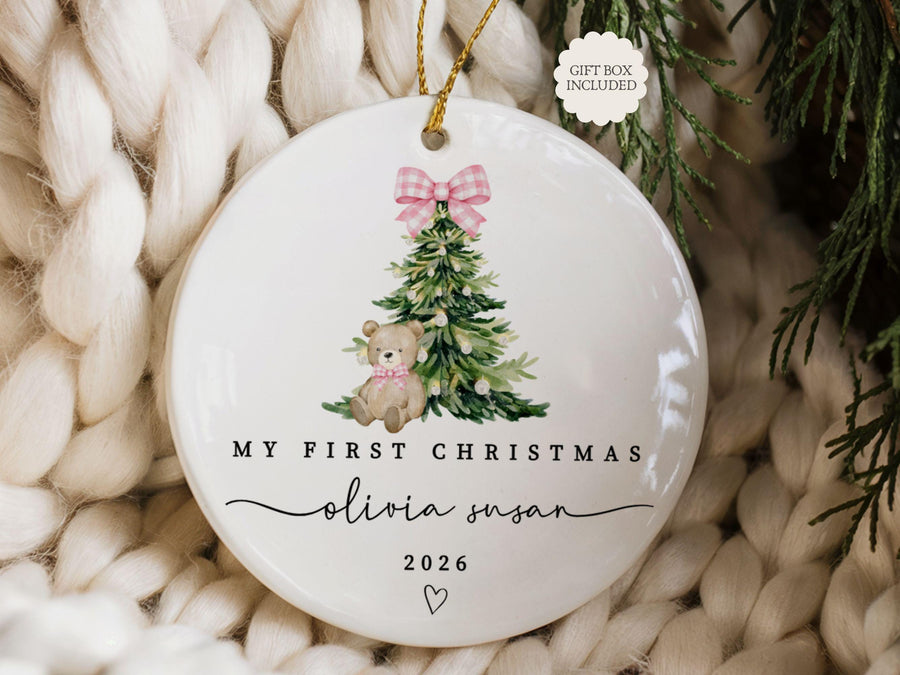 May include: A round white ceramic ornament featuring a watercolor illustration of a green Christmas tree with a pink gingham bow, a teddy bear, and the text "MY FIRST CHRISTMAS", "olivia susan", "2026", and a small heart, with a gold hanging string.