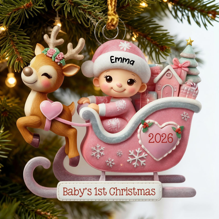 May include: A Christmas ornament featuring a reindeer with a floral crown pulling a pink sleigh. Inside the sleigh sits a baby wearing a pink hat with the name Emma. The sleigh displays a heart with 2026 and a sign reading Baby's 1st Christmas.