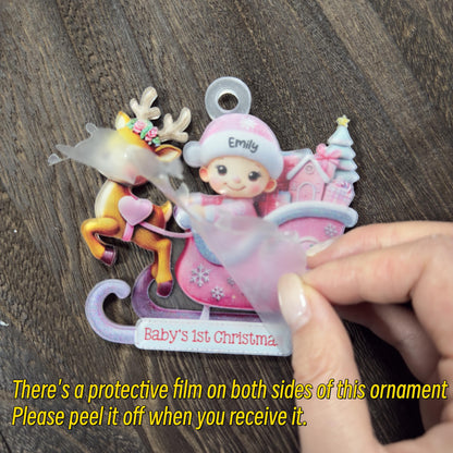 May include: A flat acrylic Christmas ornament featuring a cartoon reindeer and a child in a pink hat sitting in a sleigh with the text Emily and Baby's 1st Christmas. A translucent protective film is being peeled off the surface of the ornament.