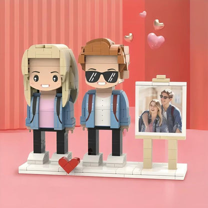 A custom building block set featuring two stylized figures, one with blonde hair and one with brown hair wearing sunglasses, standing on a white base next to a framed photograph of a couple, accented by small floating heart shapes.