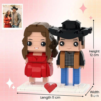 A custom brick-built figurine set featuring two characters on a white base with a red heart. The female figure has brown hair and a red dress. The male figure wears a black cowboy hat, tan vest, and blue pants. Dimensions are 12 cm high, 11 cm long, 5 cm wide.