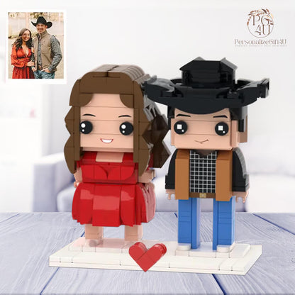 A custom building block figurine set featuring two characters on a white base with a small red heart. One character has long brown hair and a red dress, while the other wears a black cowboy hat, tan vest, and blue pants.
