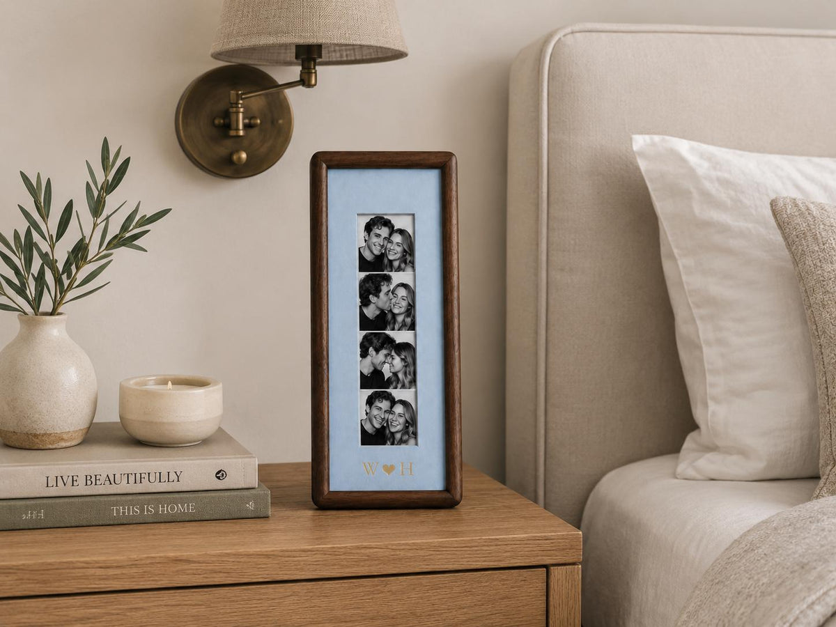 A vertical rectangular dark wood photo frame with a light blue mat board. The frame displays a strip of four black and white photographs of a couple. Below the photos, the letters W and H are separated by a small heart symbol.