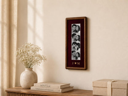 A rectangular dark wood picture frame containing a vertical strip of four black and white photographs of a couple. Below the photos, the letters J and M are separated by a heart symbol in gold lettering on a deep burgundy background.
