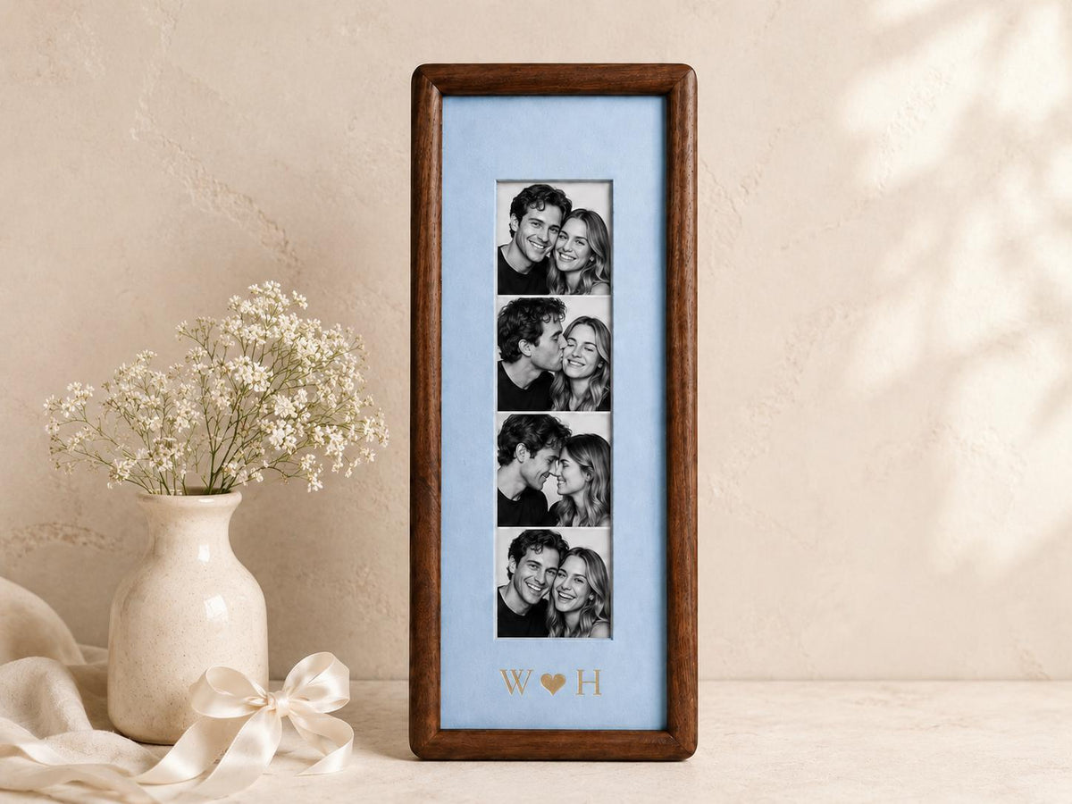 A vertical rectangular dark wood picture frame containing a light blue mat board with four stacked black and white photographs of a couple. The mat board features the gold-colored letters W and H separated by a heart symbol.