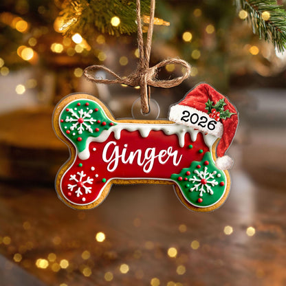 May include: A bone-shaped Christmas ornament featuring a red center with the name Ginger in white script, green ends with white snowflake icing, white icing drips, and a red Santa hat with the year 2026, all suspended by a brown twine loop.