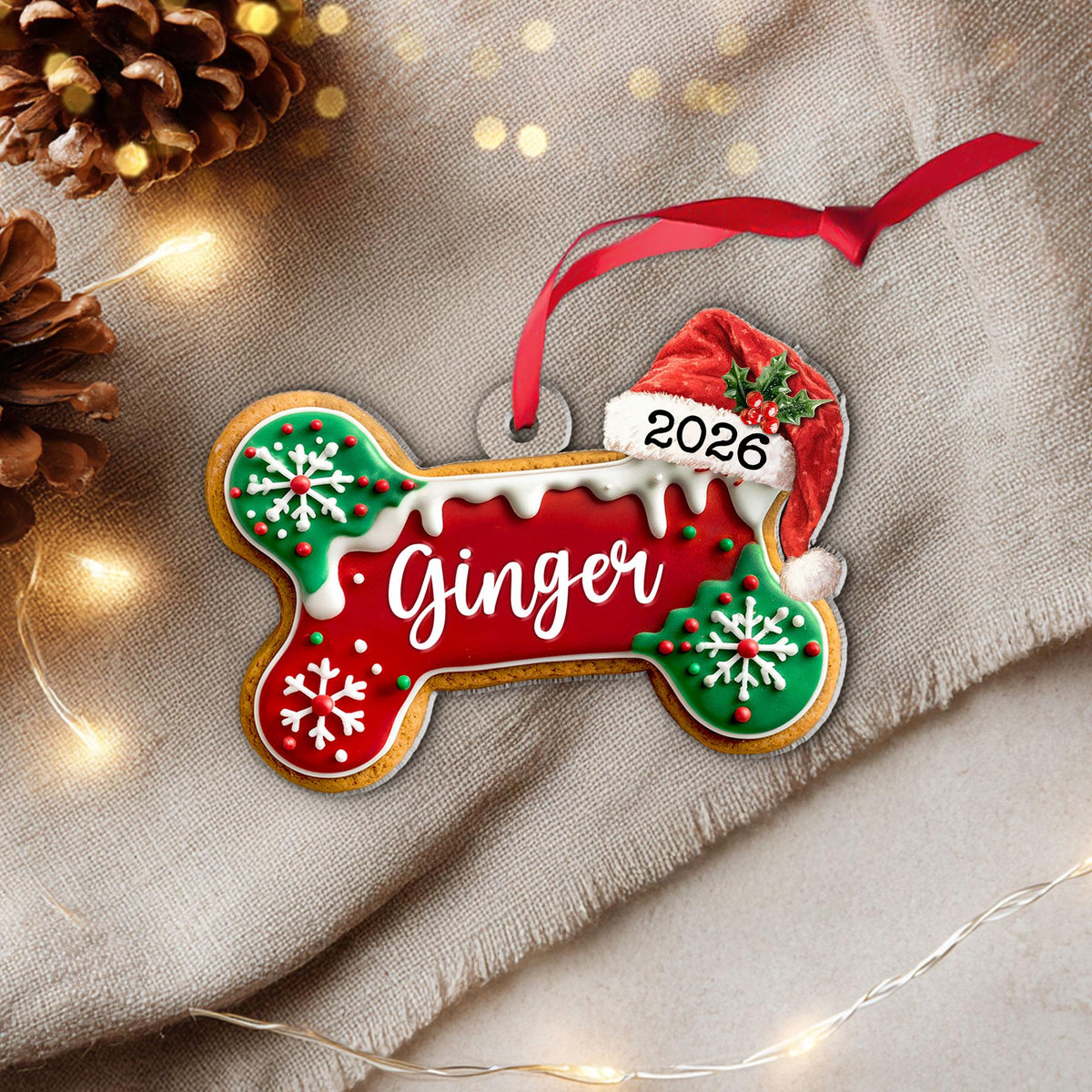 May include: A bone-shaped Christmas ornament featuring a red center with the name Ginger in white script, green ends with white snowflake icing, a Santa hat with the year 2026, and a red ribbon for hanging. The design mimics a frosted cookie.
