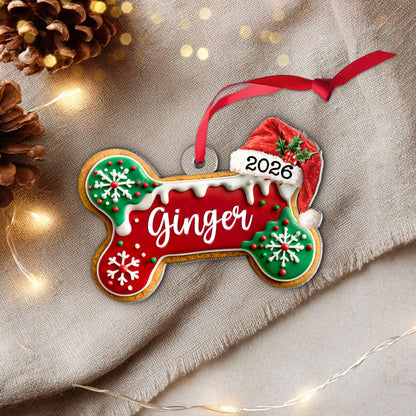 May include: A bone-shaped Christmas ornament featuring a red center with the name Ginger in white script, green ends with white snowflake icing, a Santa hat with the year 2026, and a red ribbon for hanging. The design mimics a frosted cookie.