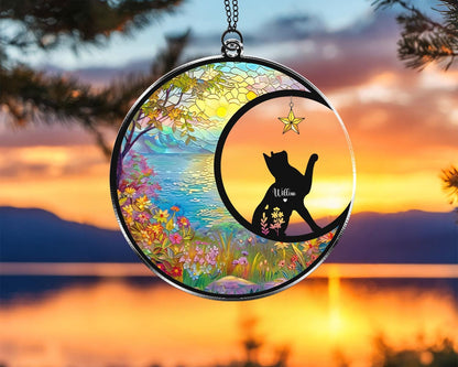 May include: A round, stained-glass suncatcher with a black cat silhouette inside a crescent moon. The cat reaches for a star. The background features a colorful landscape with flowers, water, and a sunset. The cat's name, Willow, is written on its chest.
