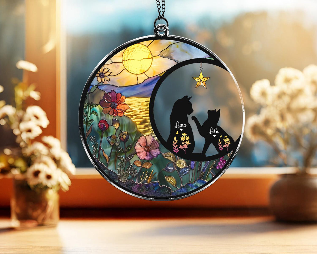 May include: A round, stained-glass suncatcher with a black metal frame. The design features a landscape with flowers, a sun, and two black cat silhouettes with the names "Luna" and "Lola". A gold star hangs above the cats.