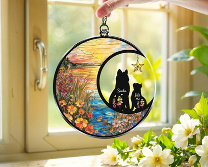 May include: A round, stained-glass suncatcher featuring a black silhouette of a dog and a cat inside a crescent moon. The names "Simba" and "Loki" are visible. The background depicts a colorful landscape with flowers and water.