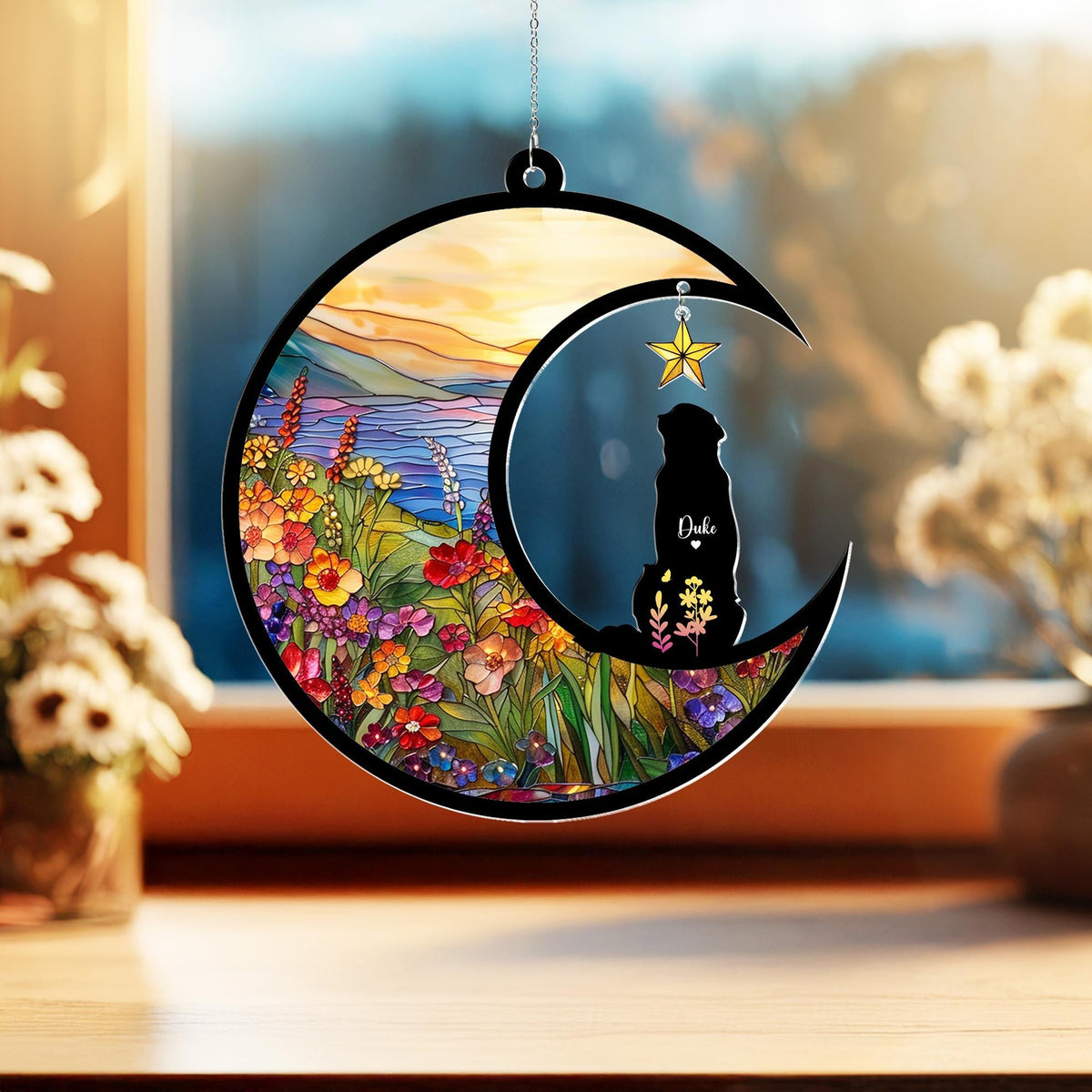 May include: A crescent moon-shaped ornament with a black border, featuring a stained-glass style landscape and a silhouette of a dog named Duke. A gold star hangs from the top. The landscape includes colorful flowers and a sunset.