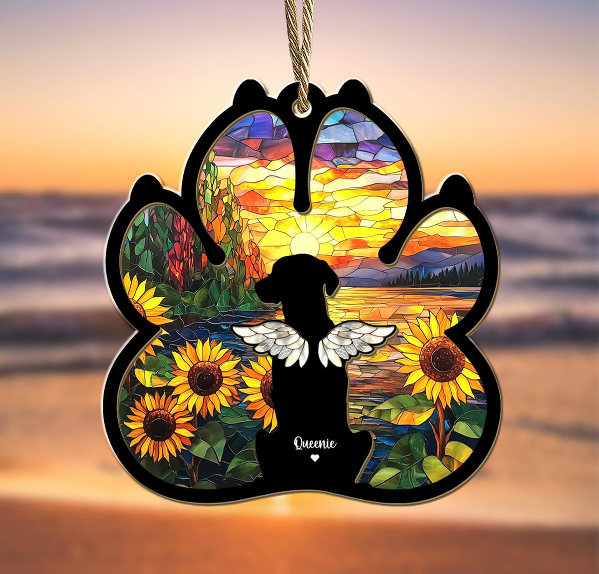 May include: A stained glass paw print ornament with a silhouette of a dog with wings in front of a sunset over a lake. The ornament has sunflowers and the name "Queenie" at the bottom.