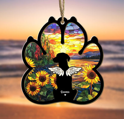 May include: A stained glass paw print ornament with a silhouette of a dog with wings in front of a sunset over a lake. The ornament has sunflowers and the name "Queenie" at the bottom.