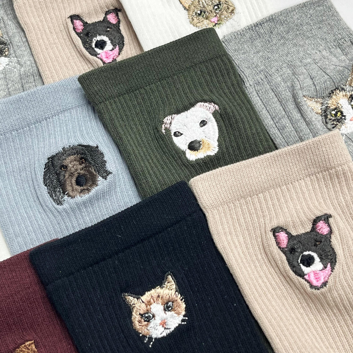 A collection of ribbed socks in various colors including beige, olive green, light blue, black, and gray. Each sock features a detailed embroidered portrait of a dog or cat face, showcasing different breeds and expressions in thread.