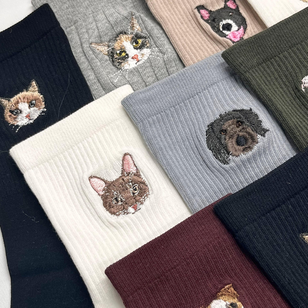 A collection of ribbed socks in various colors including black, white, gray, beige, olive green, and burgundy. Each sock features a detailed embroidered portrait of a cat or dog face, showcasing different breeds and fur patterns.
