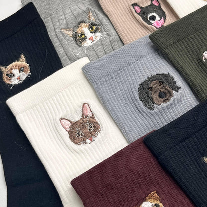 A collection of ribbed socks in various colors including black, white, gray, beige, olive green, and burgundy. Each sock features a detailed embroidered portrait of a cat or dog face, showcasing different breeds and fur patterns.