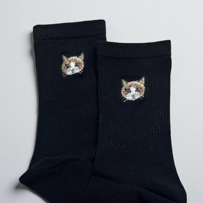 A pair of black crew socks featuring a small, detailed embroidered patch of a brown and white cat face on the upper ribbed section of each sock. The embroidery depicts the cat with pointed ears, whiskers, and a white muzzle.