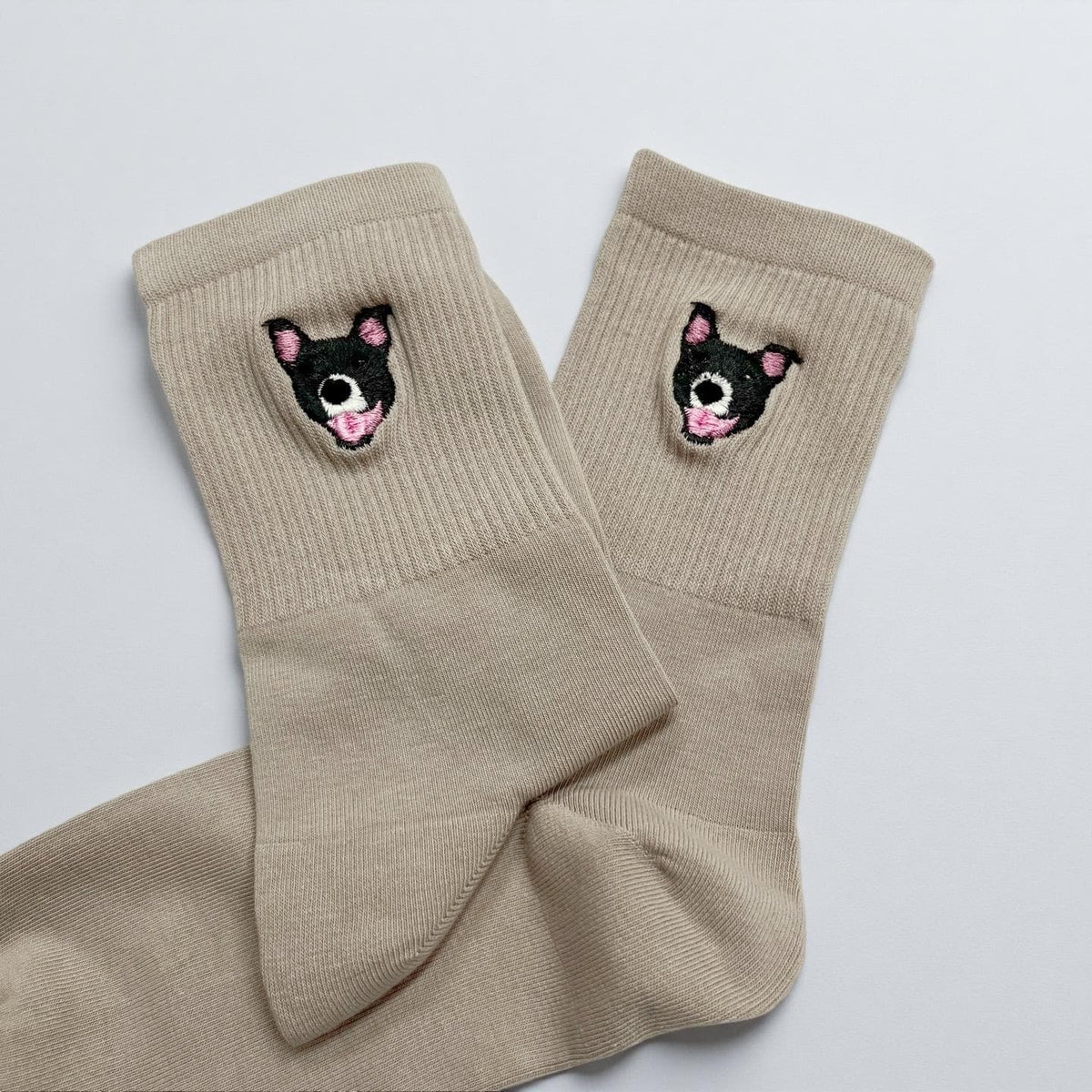 A pair of beige crew socks featuring a small embroidered graphic of a black and white dog face with pink ears and a pink tongue on the ribbed cuff of each sock. The socks are made of a solid light tan fabric with a ribbed texture.