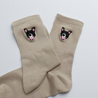 A pair of beige crew socks featuring a small embroidered graphic of a black and white dog face with pink ears and a pink tongue on the ribbed cuff of each sock. The socks are made of a solid light tan fabric with a ribbed texture.