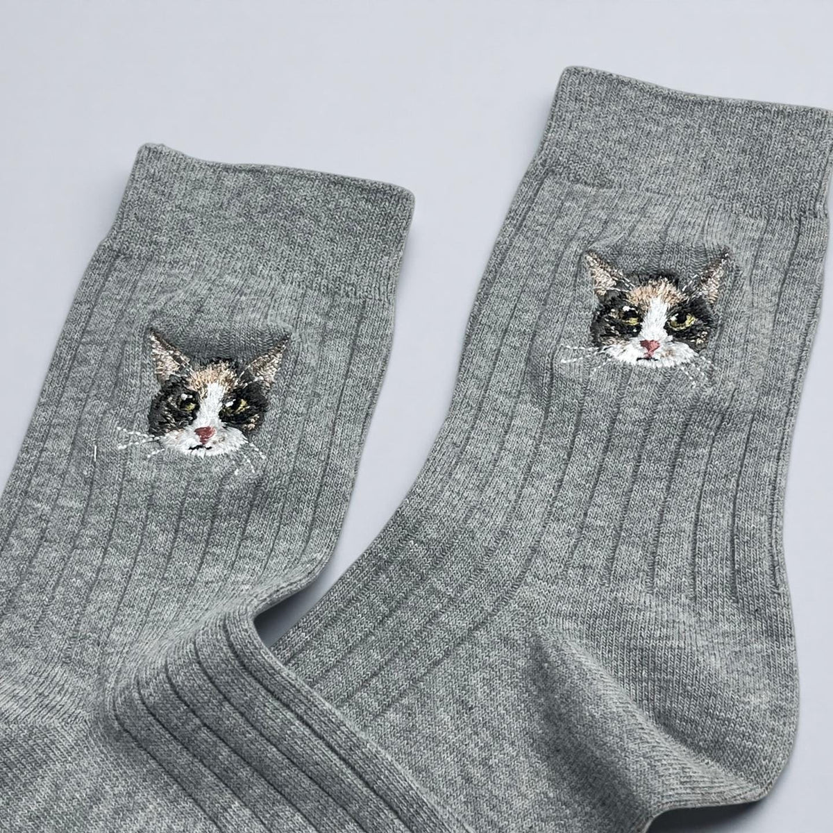 A pair of grey ribbed socks featuring an embroidered calico cat face on the upper section of each sock. The cat embroidery displays black, white, and orange fur details with white whiskers, positioned centrally on the ribbed fabric.