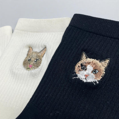 A pair of ribbed socks, one in white and one in black, each featuring a detailed embroidered cat face. The white sock displays a brown cat with a pink nose, while the black sock shows a calico cat with white, brown, and black fur.