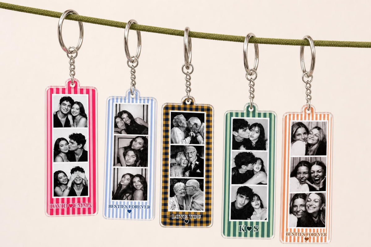 Five rectangular acrylic keychains hanging from a green cord. Each keychain features a vertical strip of three black and white photos and a colored border with stripes. Text at the bottom includes names or phrases like DAVID & EMMA.