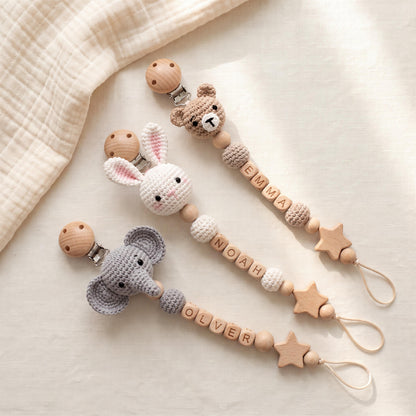 Three pacifier clips featuring crocheted animal heads: a brown bear, a white rabbit, and a gray elephant. Each clip includes wooden beads, letter beads spelling EMMA, NOAH, or OLVER, and a wooden star charm with a loop at the end.