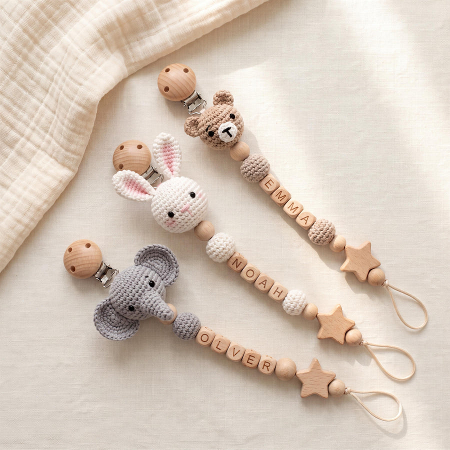 Three pacifier clips featuring crocheted animal heads: a brown bear, a white rabbit, and a gray elephant. Each clip includes wooden beads, letter beads spelling EMMA, NOAH, or OLVER, and a wooden star charm with a loop at the end.