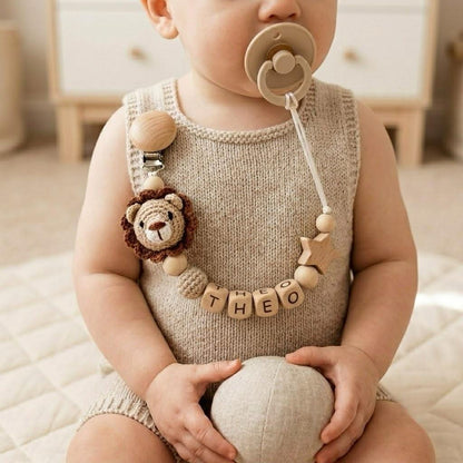 A beige pacifier clip featuring a crocheted lion head, a wooden star, and wooden beads spelling the name THEO. The clip is attached to a beige knitted romper, and a beige pacifier is held in the mouth of the wearer.