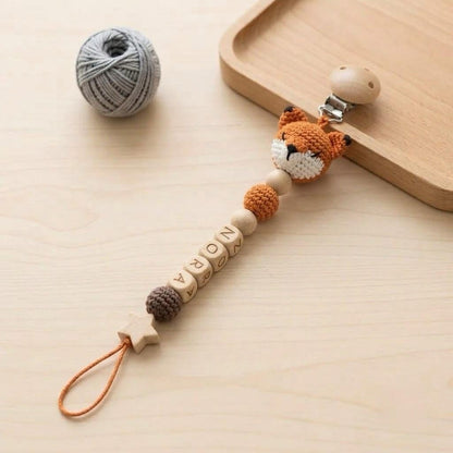 A pacifier clip featuring a crocheted orange and white fox head, wooden beads spelling the name NORA, a small brown crocheted sphere, and a wooden star-shaped bead. The clip includes a metal fastener and an orange cord loop.