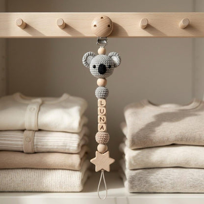A pacifier clip featuring a crocheted gray koala head, wooden beads, and four square wooden beads spelling the name LUNA. The clip includes a light wood star-shaped charm at the bottom and a loop for attachment, hanging from a wooden rack.