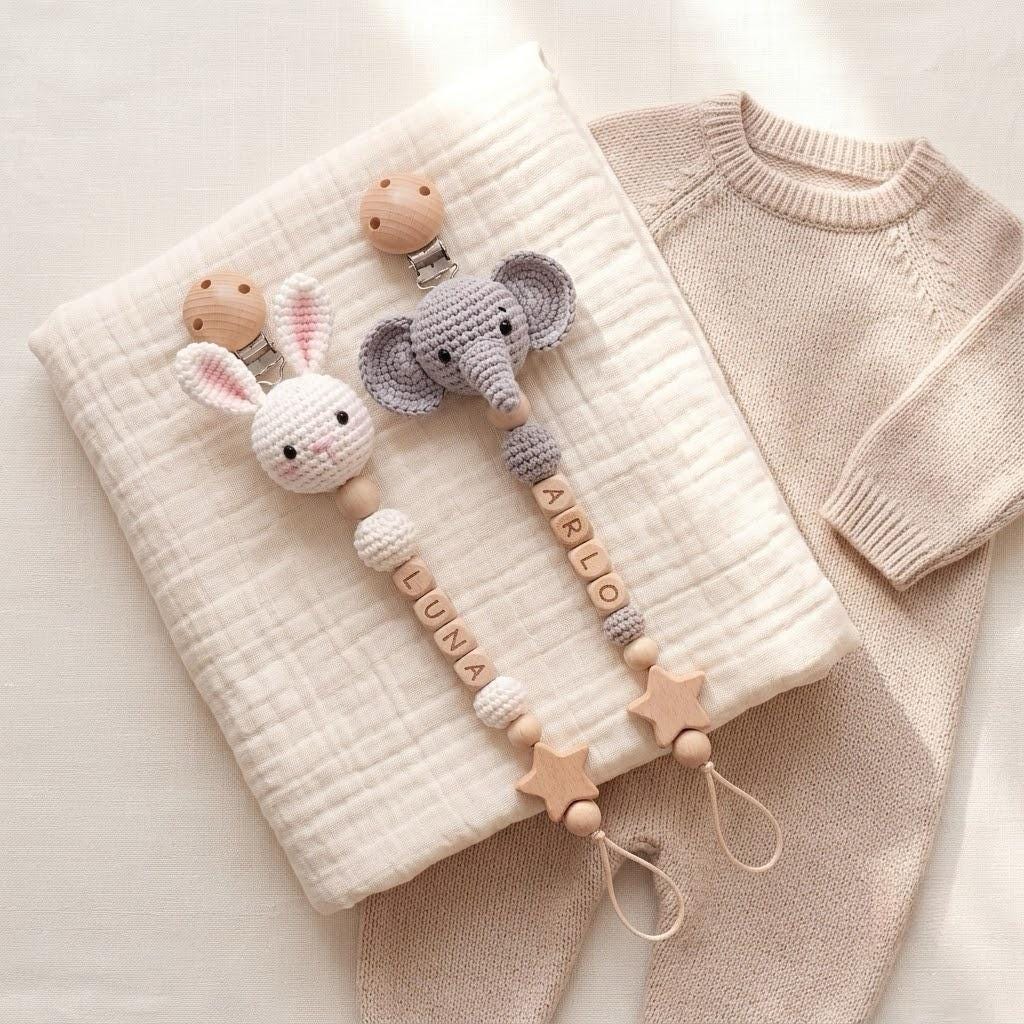 Two pacifier clips featuring crocheted animal heads, one a white rabbit and one a gray elephant. Each clip includes wooden beads, some engraved with the names LUNA and ARLO, and wooden star-shaped accents with a loop at the bottom.