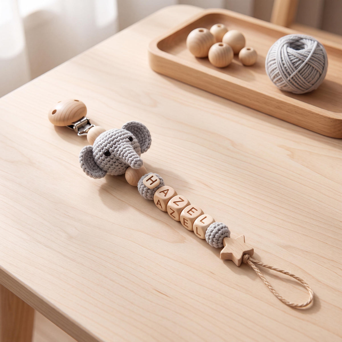 A pacifier clip featuring a crocheted gray elephant head, natural wooden beads, and light wooden cube beads spelling the name HAZEL. The clip includes a star-shaped wooden charm at the end with a looped cord for attachment.