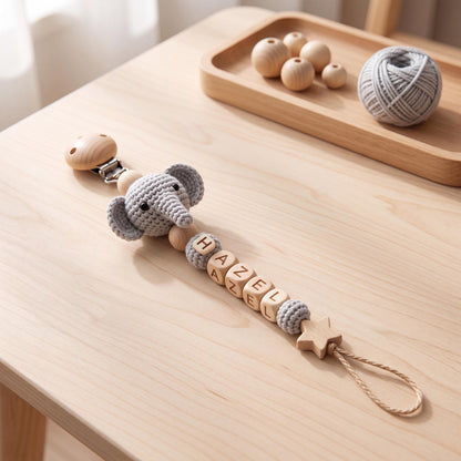 A pacifier clip featuring a crocheted gray elephant head, natural wooden beads, and light wooden cube beads spelling the name HAZEL. The clip includes a star-shaped wooden charm at the end with a looped cord for attachment.
