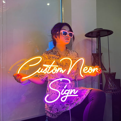 A neon sign with the text "Custom Neon Sign" in a cursive font. The sign is lit up in a warm yellow and pink color. A person is holding the sign in front of them.