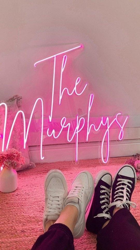 Pink neon sign with the text "The Murphys" in a cursive font. The sign is hanging on a pink wall. Two pairs of shoes are in the foreground, one pair is white sneakers and the other pair is black high-top sneakers.