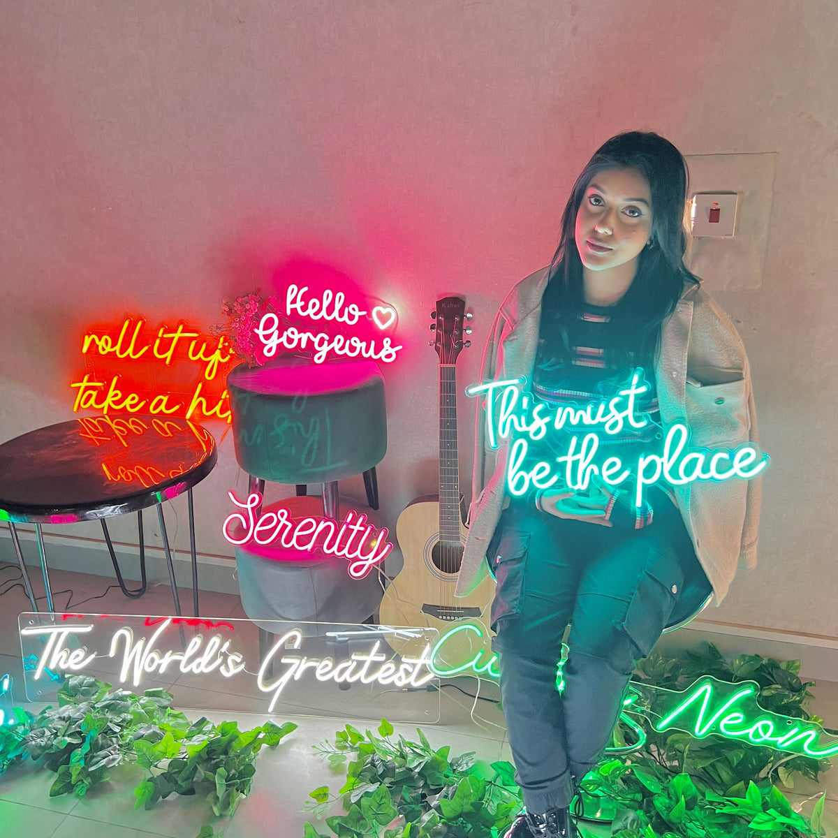 A woman poses in front of a wall with neon signs. The signs read "Hello Gorgeous", "Roll it up, take a hit", "Serenity", "This must be the place", "The World's Greatest", and "Neon".