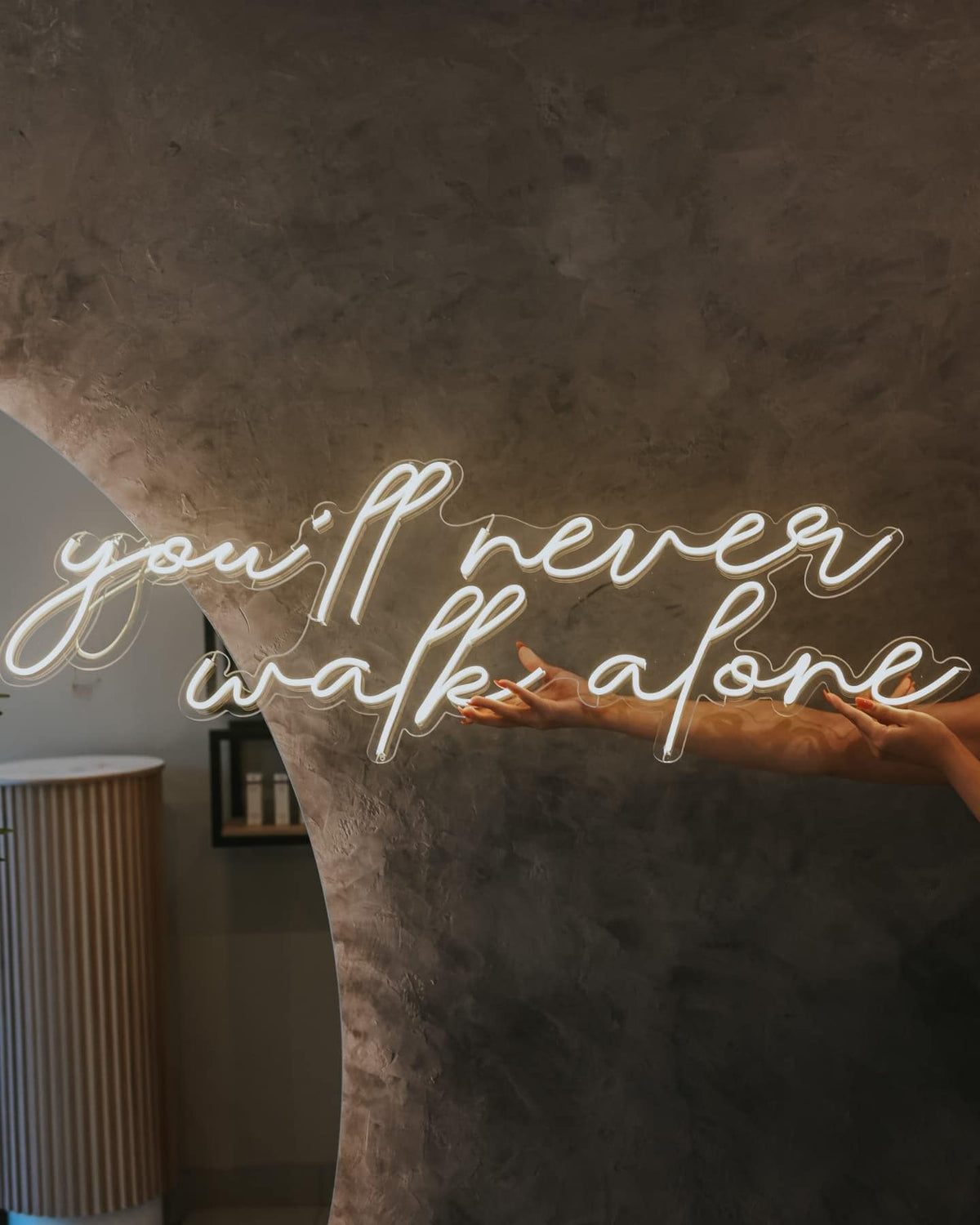 White neon sign with the text "you'll never walk alone" on a brown textured wall.