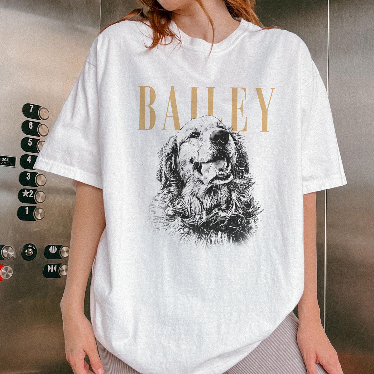White t-shirt featuring a detailed black and white illustration of a Golden Retriever dog with its tongue out. The word "BAILEY" is printed in gold above the dog image. The shirt has short sleeves.
