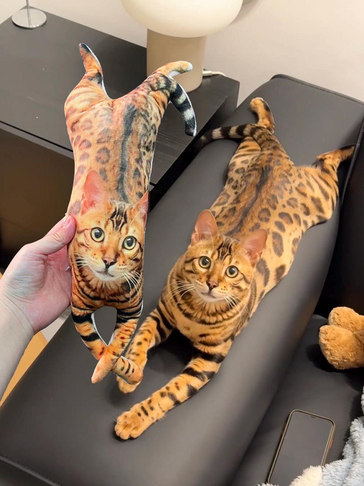 A plush toy shaped like a Bengal cat with a realistic brown and black spotted coat pattern, featuring a printed face with large eyes, held next to a live Bengal cat with an identical coat pattern and markings lying down.