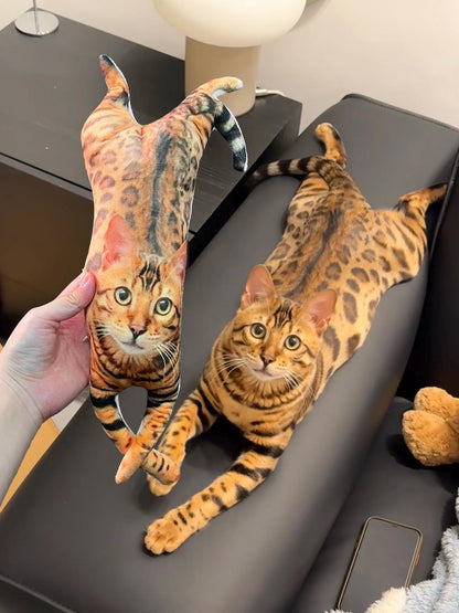 A plush toy shaped like a Bengal cat with a realistic brown and black spotted coat pattern, featuring a printed face with large eyes, held next to a live Bengal cat with an identical coat pattern and markings lying down.