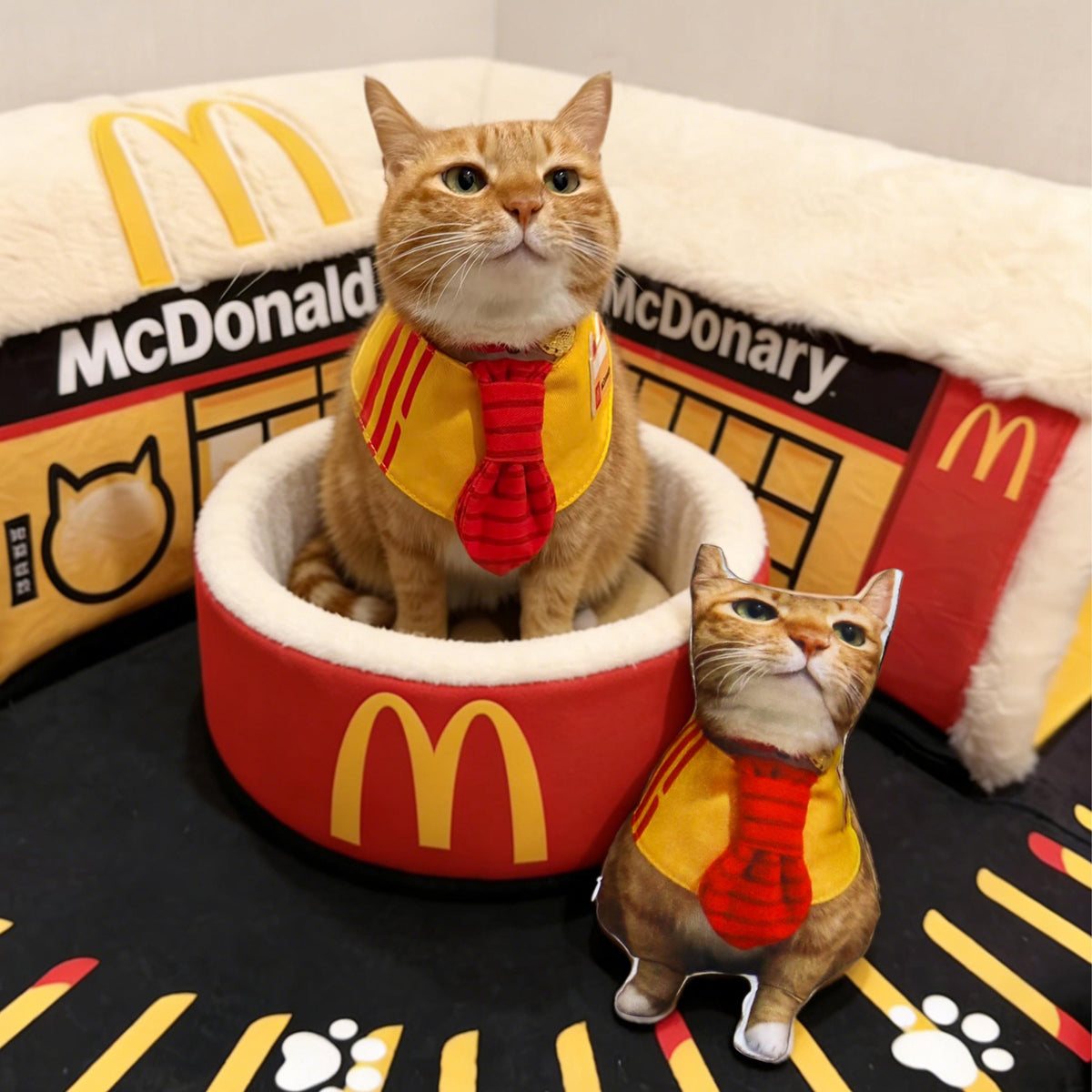 An orange tabby cat wearing a yellow bandana with a red tie sits in a round red pet bed featuring the yellow McDonald's logo. Beside the bed is a small plush toy shaped like a cat wearing an identical yellow bandana and red tie.