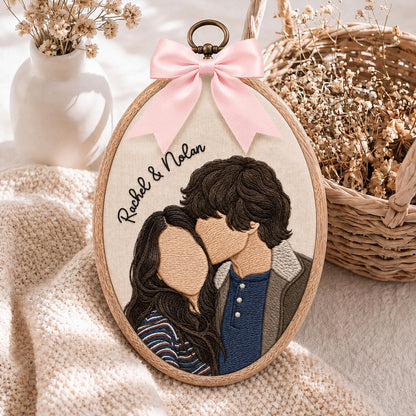 An oval embroidery hoop featuring a custom thread portrait of two people with the text Rachel & Nolan stitched in black. The hoop is adorned with a light pink satin ribbon bow at the top and a bronze-colored metal hanging loop.