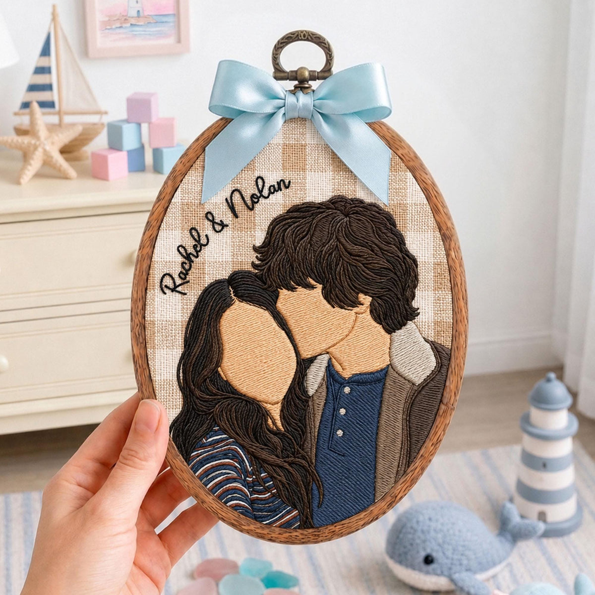 An oval embroidery hoop featuring a stitched portrait of two people with brown hair. The fabric background is a beige and white gingham pattern. The text "Rachel & Nolan" is embroidered in black. A light blue satin bow is attached at the top.