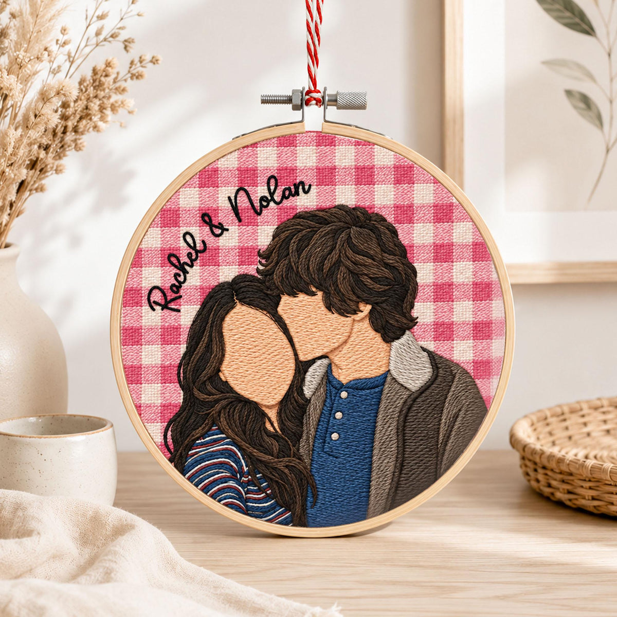 A circular embroidery hoop featuring a stitched portrait of two people against a pink and white gingham background. The text "Rachel & Nolan" is embroidered in black script. The hoop is suspended by a red and white twisted cord.