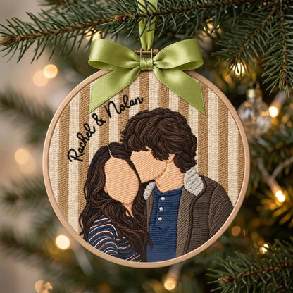 A circular embroidery hoop ornament featuring a stitched portrait of two people with brown hair, one wearing a blue shirt and brown jacket, the other a striped top. The text "Rachel & Nolan" is embroidered above, topped with a green ribbon.